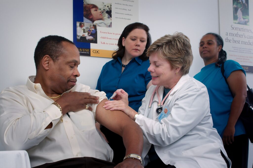 A white nurse administers an injection to a Black masculine person while two feminine people wearing blue wait behind
