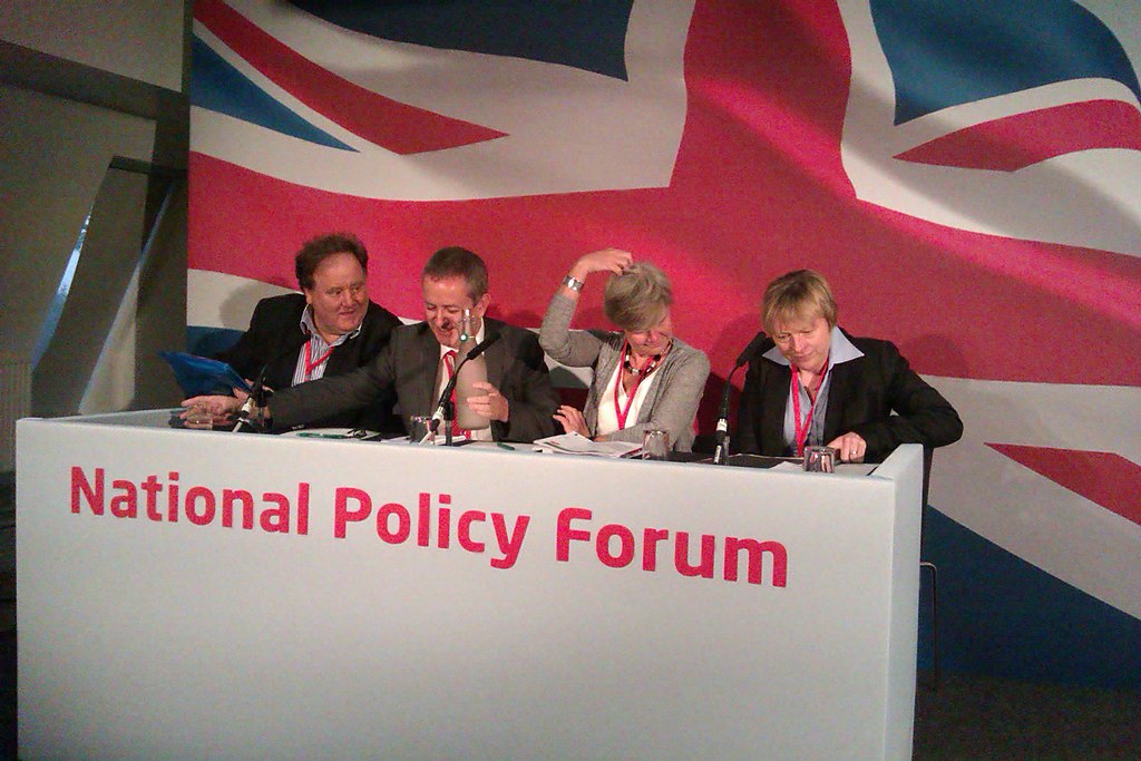 Four white people in suits in front of a British flag behind a desk labelled "National Policy Forum"
