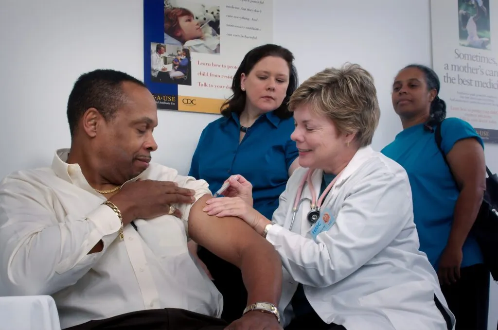 A white nurse administers an injection to a Black masculine person while two feminine people wearing blue wait behind