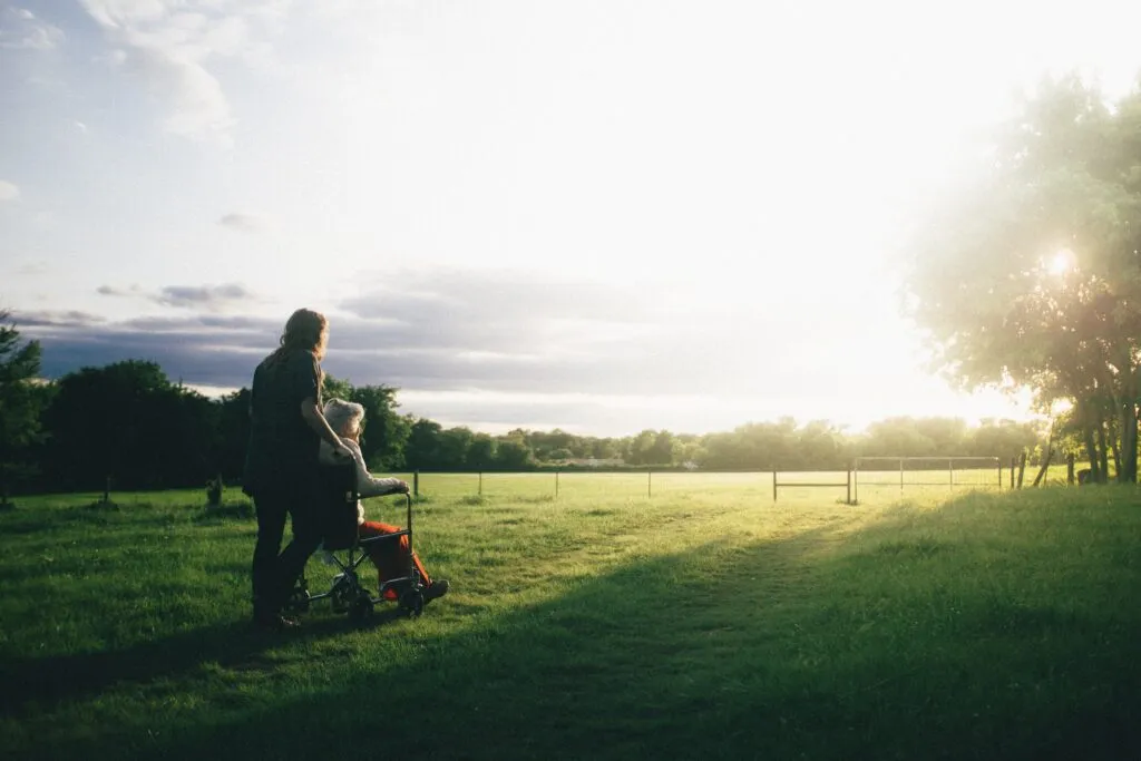 A person in a wheelchair being taken across a sunlit grassy field by a person with long hair